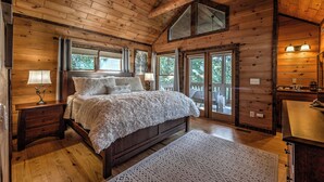 Une chambre de style cabane en bois, avec un grand lit, deux lampes de chevet, une commode en bois et une vue sur l’extérieur grâce à de grandes fenêtres.