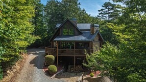Une cabane en rondins à deux étages, dotée d’une cheminée en pierre, d’un balcon et d’une allée en gravier, le tout entouré d’arbres.