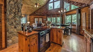 L’intérieur d’un chalet rustique, avec un mur en pierre, un plancher en bois, une grande cuisine équipée d’un poêle et de grandes fenêtres offrant une vue sur l’extérieur.