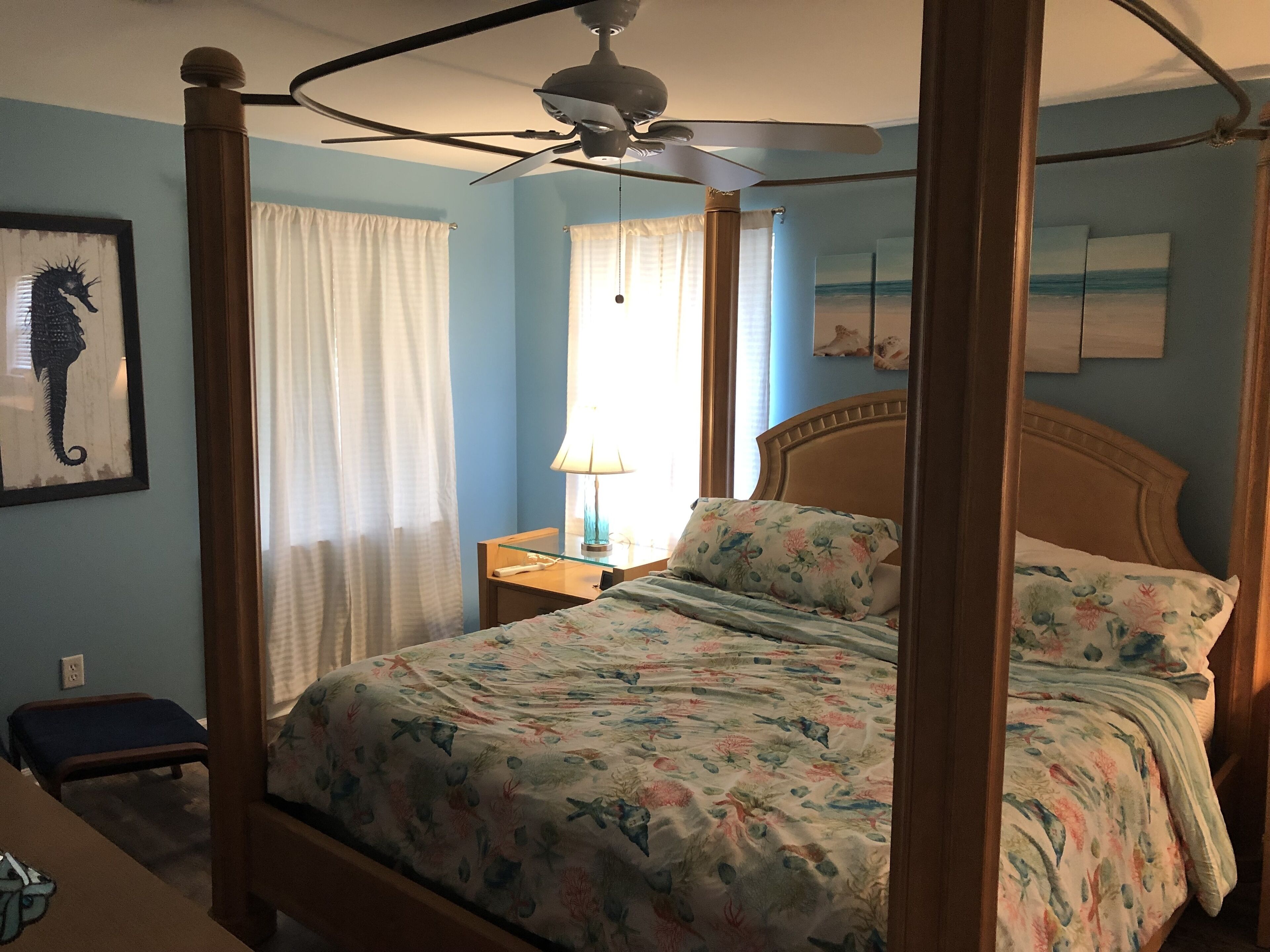 A four-poster bed with a floral bedspread, a ceiling fan, and a nightstand with a lamp.