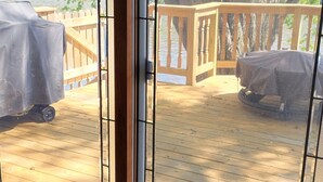 Blick durch eine Glastür auf eine Terrassenfläche mit Holzgeländer, einem Tisch und einem Stuhl, beide überdacht.