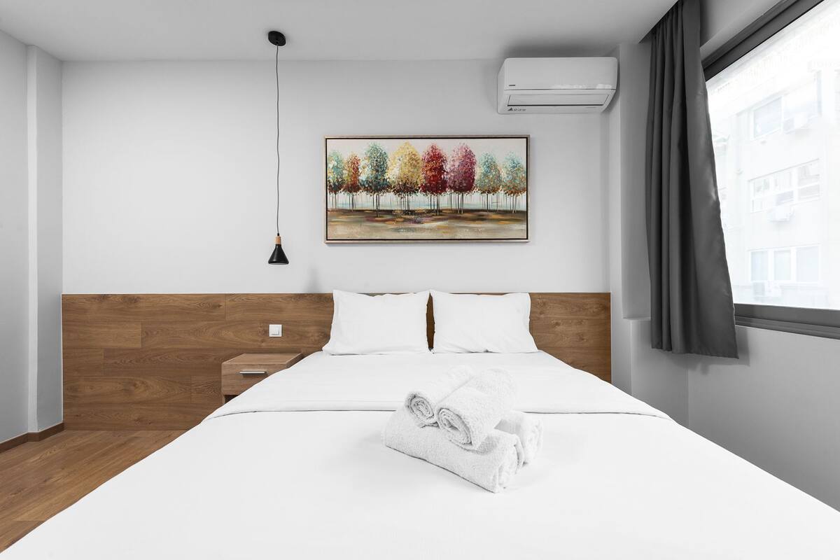 Una habitación de hotel moderna con cabecera de madera, una cama bien hecha con ropa blanca, un cuadro de árboles en la pared, una lámpara de noche y un equipo de aire acondicionado.