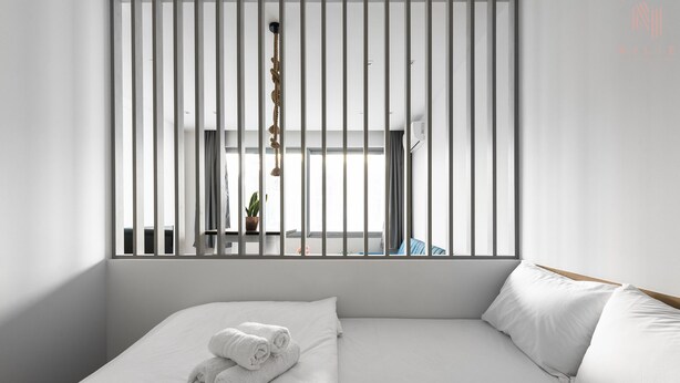 Ein modernes Hotelzimmer mit einem großen Bett, weißen Leinentextilien, Blick durch ein Fenster mit vertikalen Jalousien und einer hängenden Pflanze.