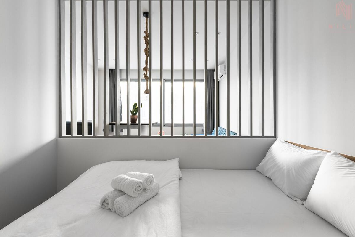 Habitación de hotel moderna con una cama grande, ropa de cama blanca, vistas a través de una ventana con persianas verticales y una planta colgante.