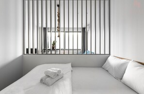 A modern hotel room with a large bed, white linens, a view through a window with vertical blinds, and a hanging plant. - Ipanema Nilie Hospitality MGMT (Thessaloniki)