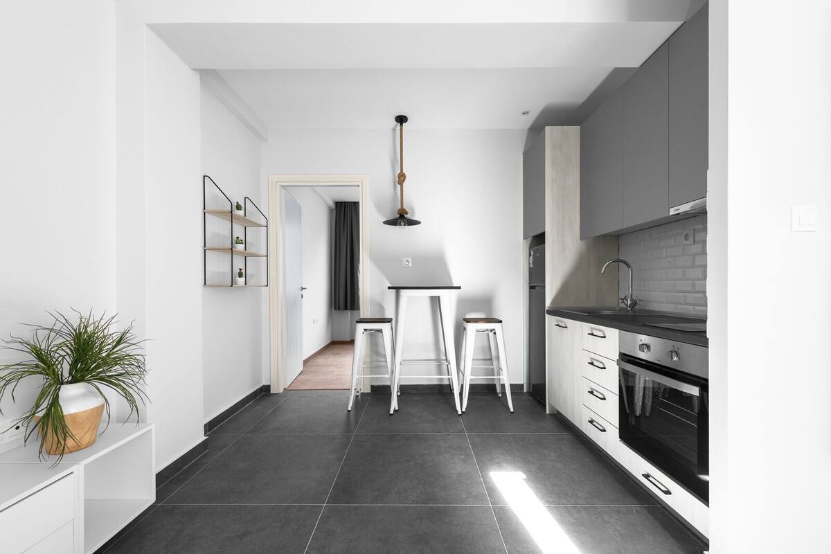 Una cocina moderna con baldosas oscuras, muebles blancos, una estufa negra y una lámpara colgante.
