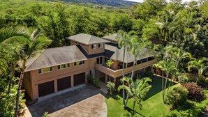 A two-story house with a garage, surrounded by lush greenery and palm trees.