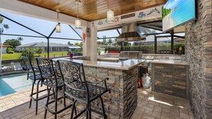 An outdoor kitchen with a stone island, bar stools, and a large TV screen.