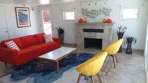 Un salón con un sofá rojo, una mesita de centro y dos sillas amarillas. Hay una chimenea con adornos encima y un cuadro en la pared.