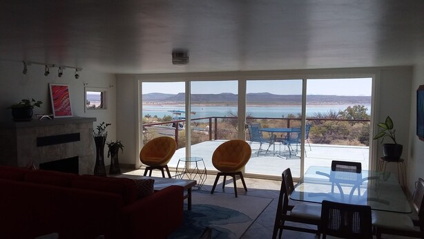 Una sala de estar con chimenea, dos sillones naranja, una mesa de vidrio y vistas a un lago y montañas a través de ventanas amplias.