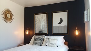 Un dormitorio con una cama, dos lámparas fijadas a la pared, un adorno decorativo con forma de sol y dos obras de arte enmarcadas que representan diseños abstractos.