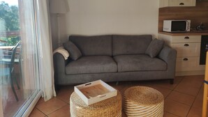 Een woonkamer met een grijze bank, een schilderij van zeilboten, een magnetron en een kleine keukenhoek.