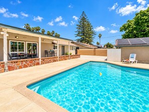 A swimming pool with a stone border, a covered patio area with outdoor furniture, and a view of a residential building with a sloped roof.