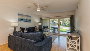 Ein modernes Wohnzimmer mit grauem Sofa, Deckenventilator und Blick ins Freie durch große Schiebeglasfenster.