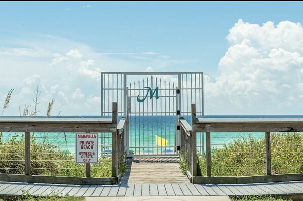 Acceso privado a una playa con una pasarela de madera que lleva a una zona vallada señalizada con una 'M' grande.