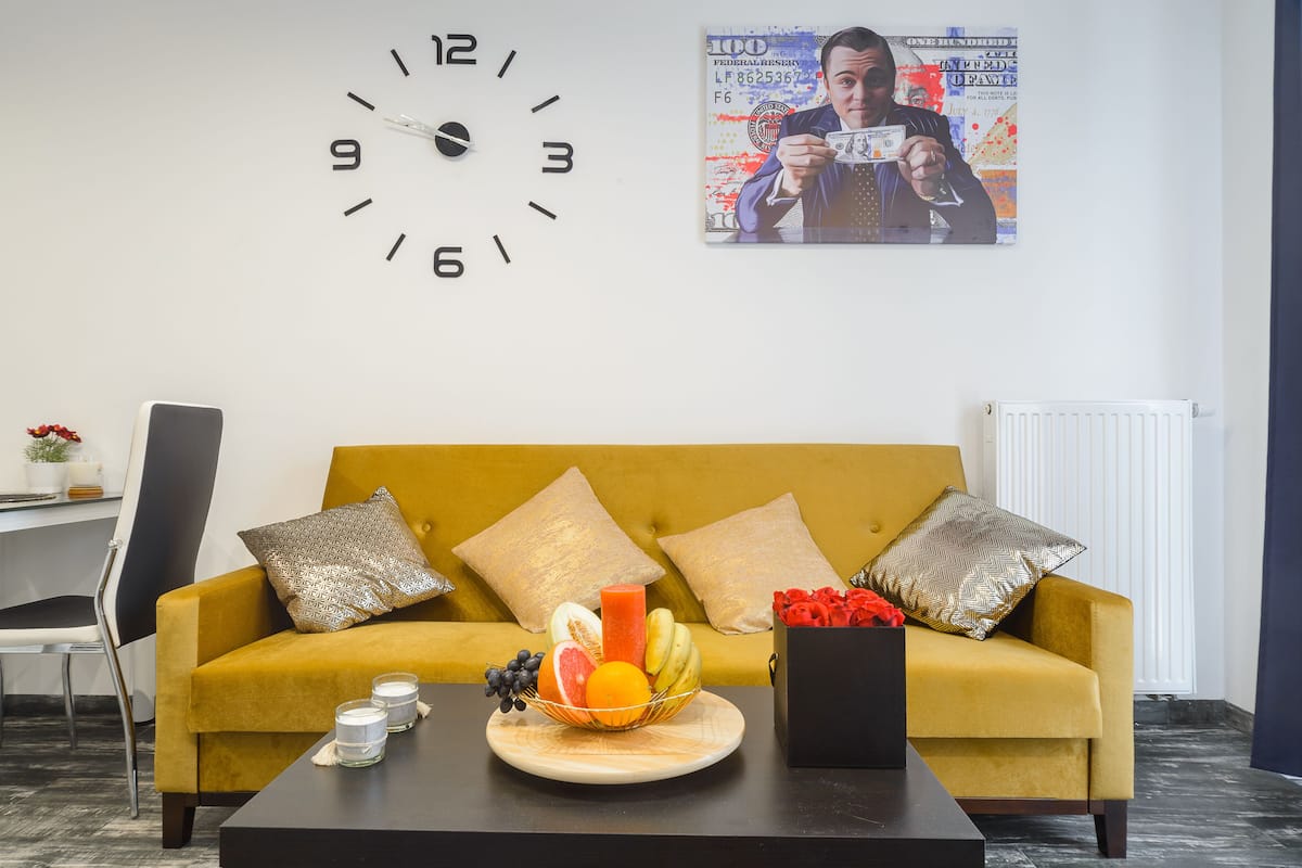 Uma sala de estar moderna com um sofá amarelo, um relógio, uma fruteira e um pôster na parede.