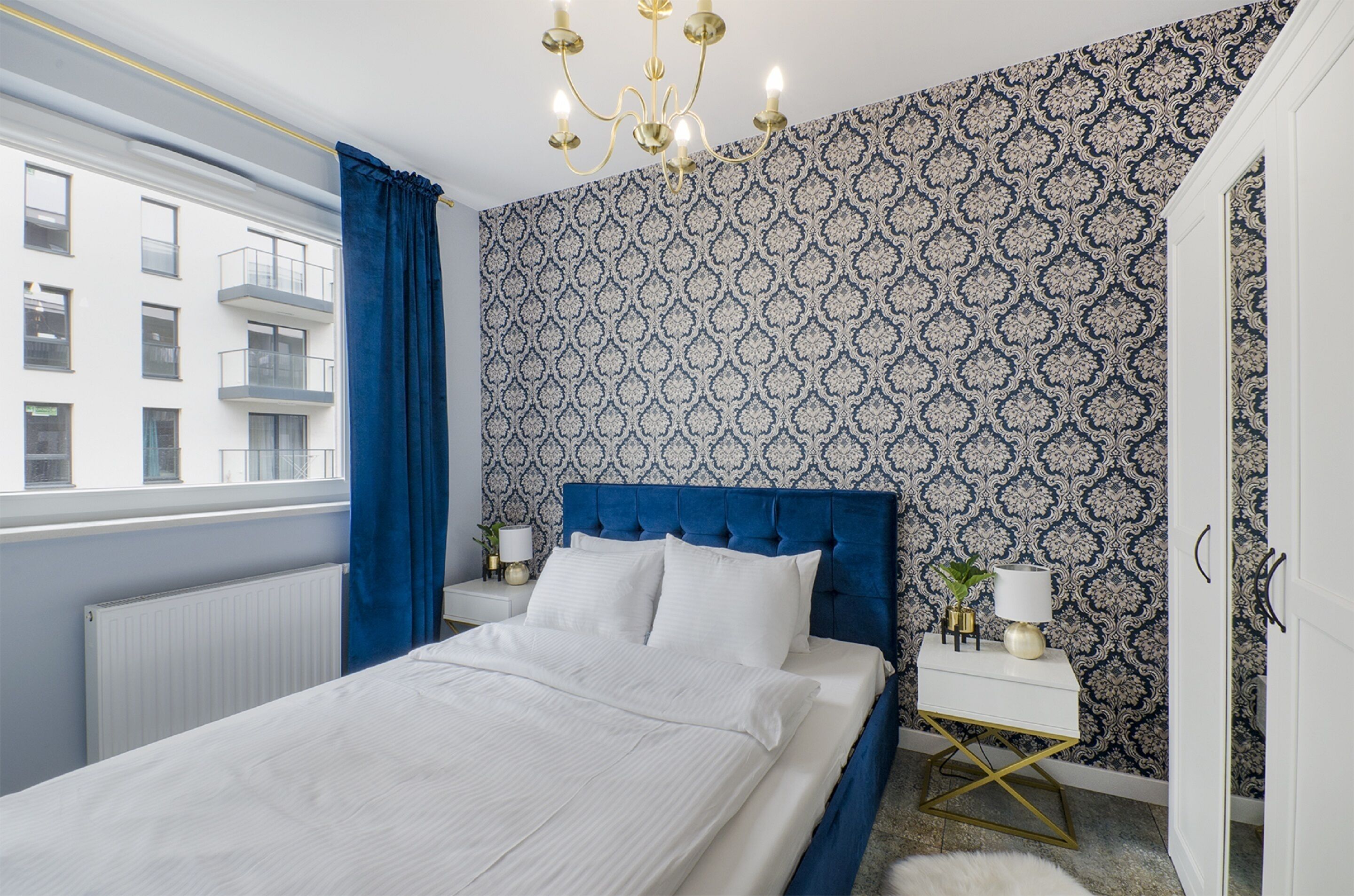 Een slaapkamer met een groot bed, een blauw hoofdeinde, een witte nachtkastje, een gouden lamp en behang met een patroon.