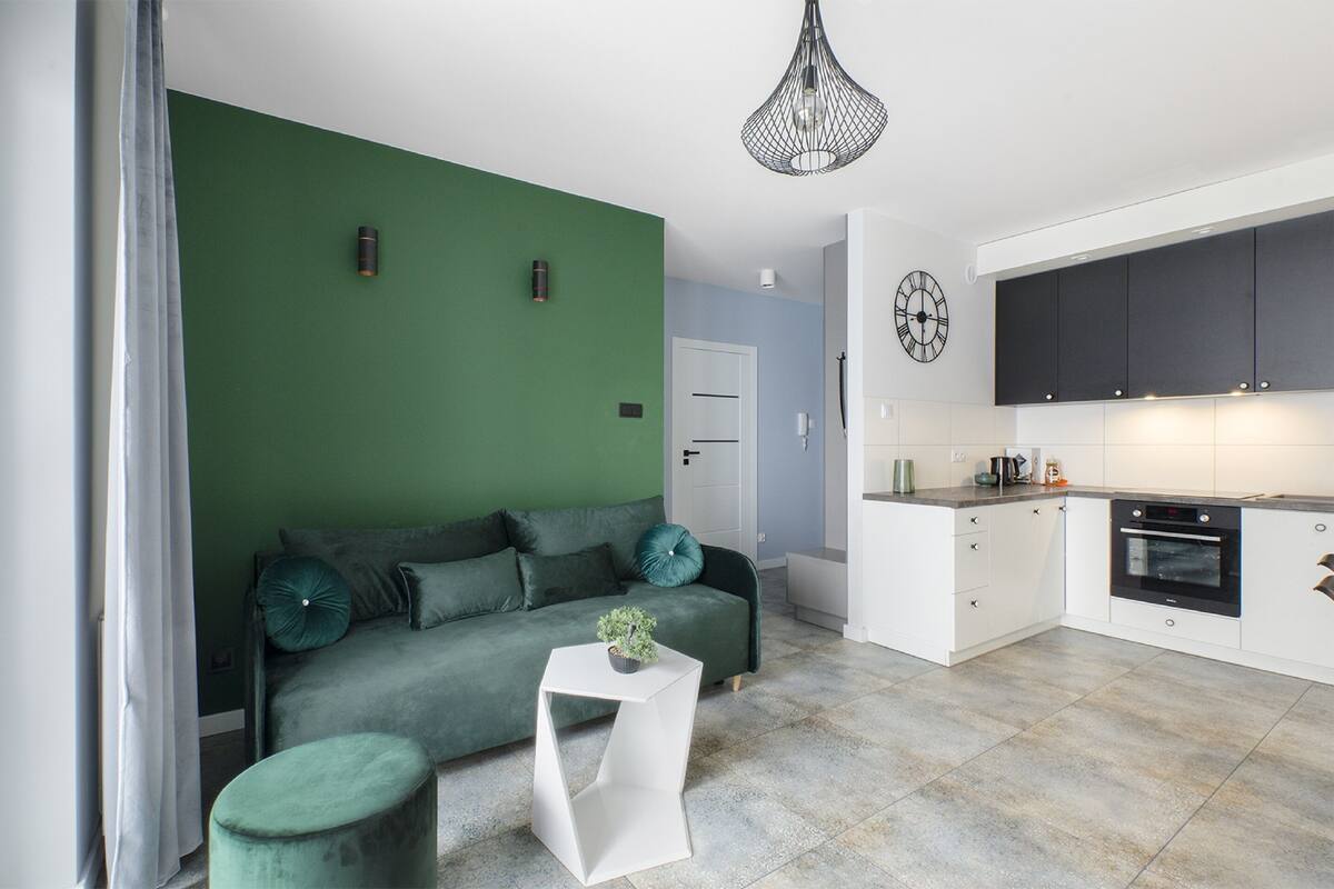 Uma sala de estar moderna com uma parede verde, um sofá, uma mesa lateral branca e uma área de cozinha com armários brancos e um forno preto.