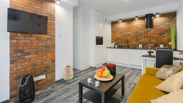 A modern living room with a flat-screen TV, a dining table with fruit and candles, a mustard-colored sofa, and a kitchen area with a brick backsplash. - Lumina apartment on Drewnowska (Lodz)