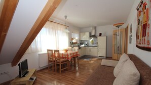 A cozy living area with a wooden floor, a brown sofa, a dining table with chairs, a kitchen in the background, and a TV on a small stand.