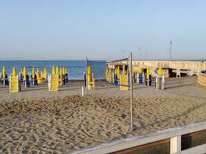 A beach with yellow and blue beach chairs and umbrellas, a wooden pier, and a concrete building with graffiti. - 500 meters from the ocean, 50 meters from the train station, rooms with all the amenities (Lido di Ostia)