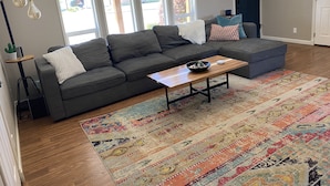 Ein Wohnzimmer mit einem grauen Ecksofa, einem Holztisch und einem farbenfrohen Teppich.