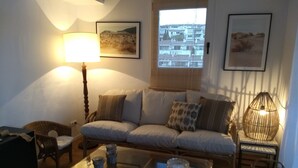 A living room with a sofa, a coffee table, a lamp, and two chairs. There are two framed pictures on the wall and a window with a view of buildings. - Casa Margot 2 (Cadaqués)