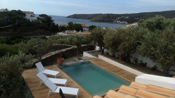 A pool area with white lounge chairs, surrounded by greenery and overlooking a coastal view. - Casa Margot 2 (Cadaqués)