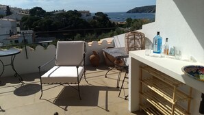 A balcony with a white chair, a wicker table, and a small bar area with bottles and glasses. - Casa Margot 2 (Cadaqués)