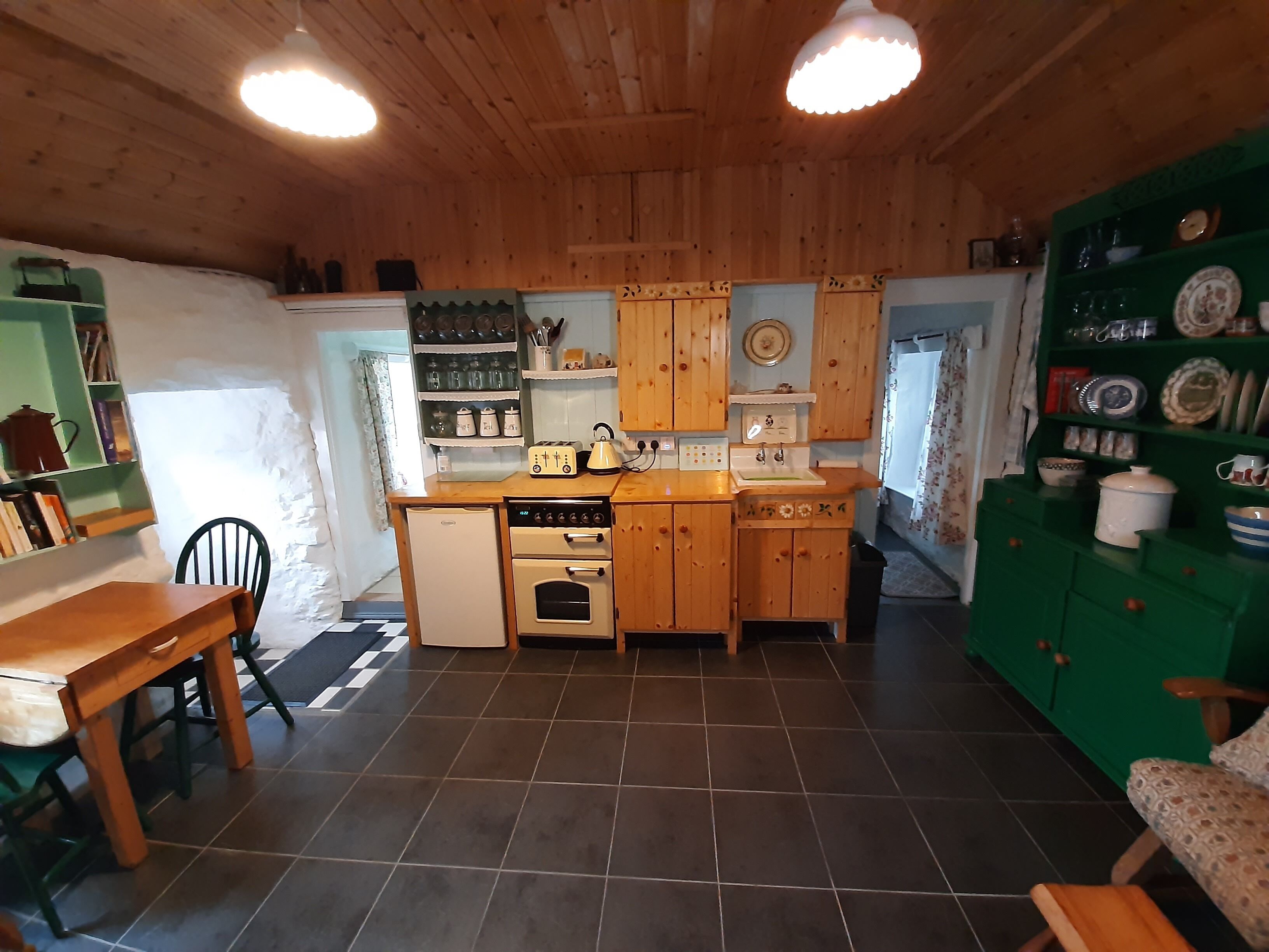 Une cuisine compacte avec des meubles en bois, un petit réfrigérateur, un évier et une cuisinière. On y trouve une table à manger avec des chaises et un meuble vert contenant de la vaisselle.