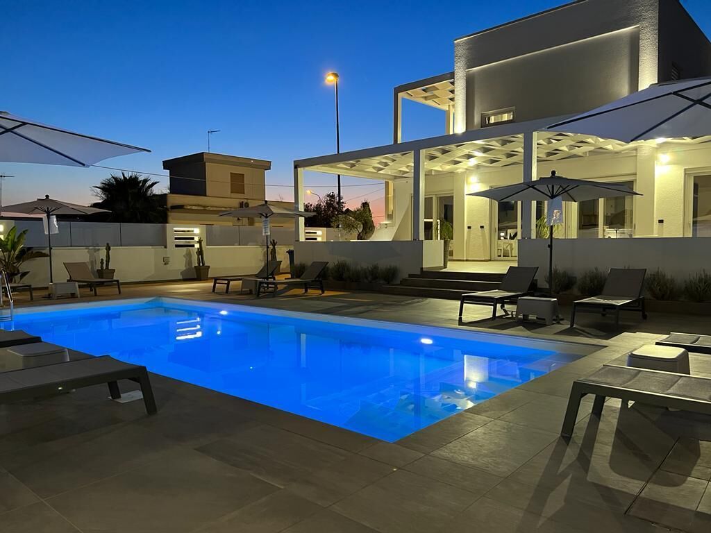 Area piscina moderna all'aperto con lettini e ombrelloni, circondata da un'abitazione contemporanea con veranda coperta.
