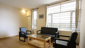 A living room with a black sofa, a glass coffee table, and two black chairs. There are large windows with blinds and a door with a picture on the wall. - The Hill Side (Ootacamund)