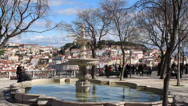 Una plaza pública con una fuente, rodeada de árboles y vistas a un paisaje urbano con edificios.