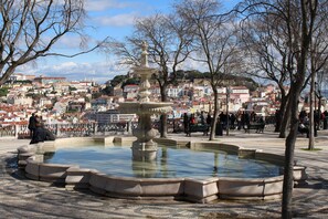 Praça pública com fonte, rodeada por árvores e vista para o horizonte urbano com edifícios.