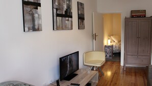 Una sala de estar con televisión de pantalla plana, una mesa de centro de madera y un sillón. Se observan pinturas abstractas en la pared y un aparador en la esquina.