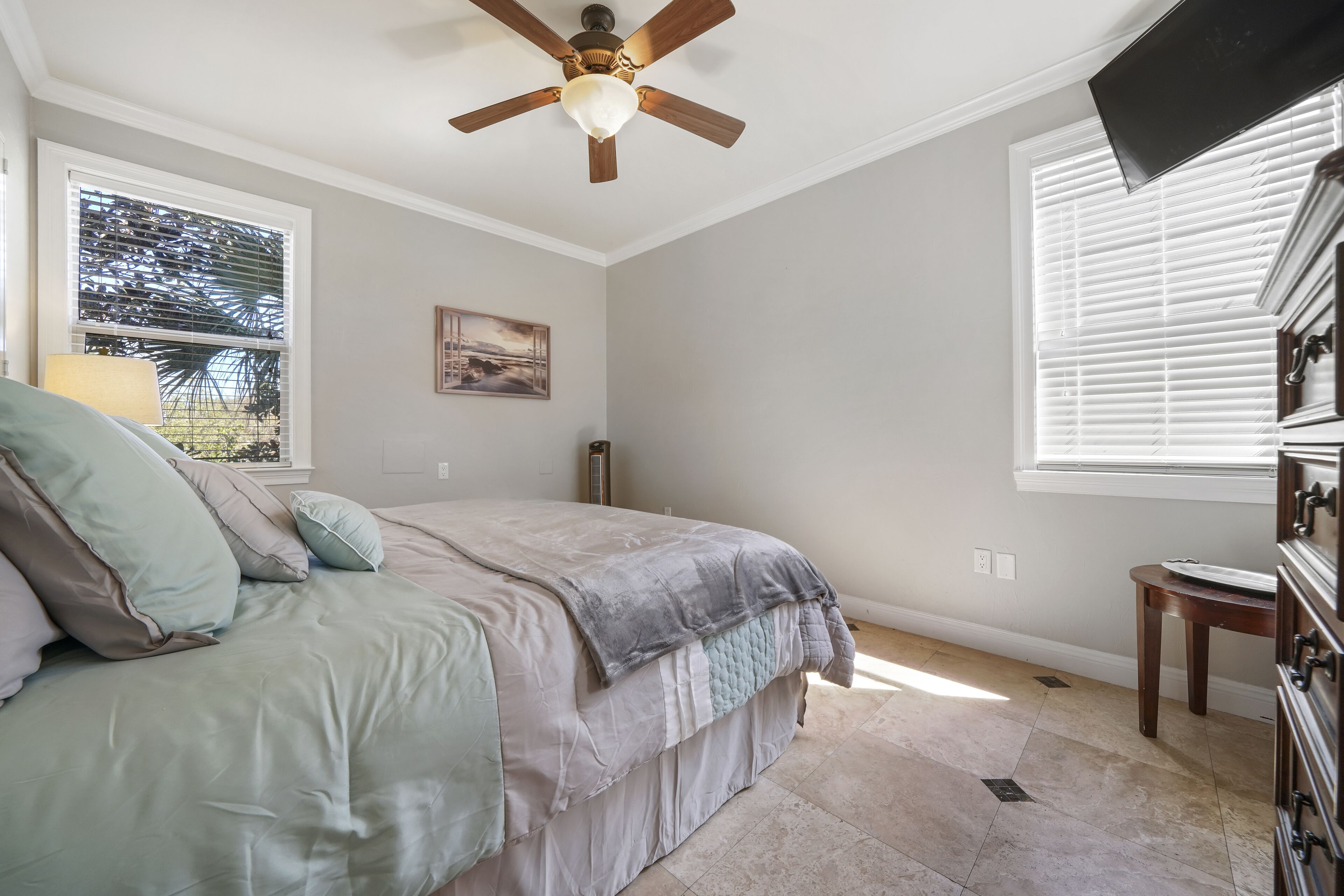A bedroom with a bed, a ceiling fan, a window with blinds, a dresser, and a small table.