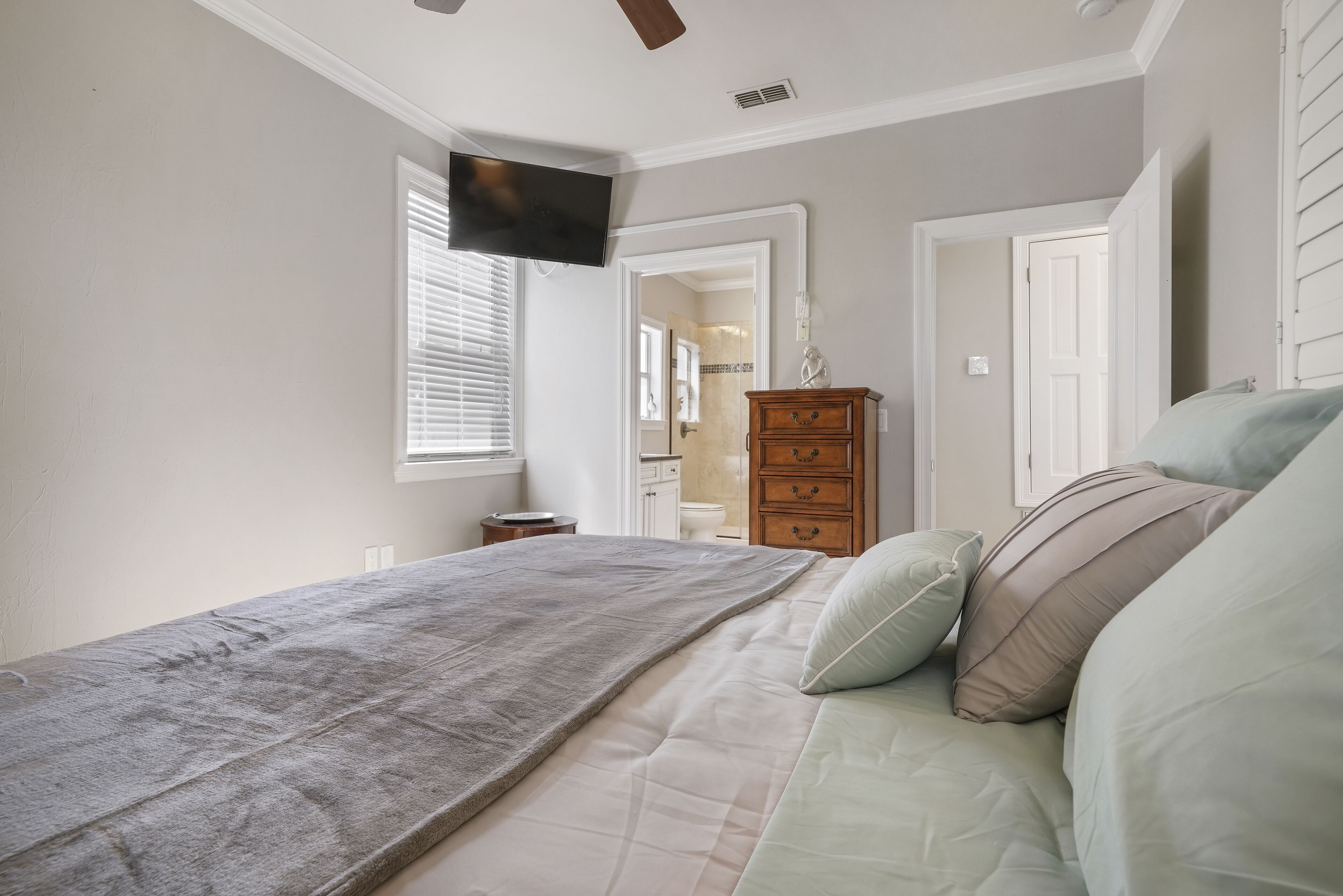 Un dormitorio con cama, cómoda, televisor montado en la pared y ventana con persianas.