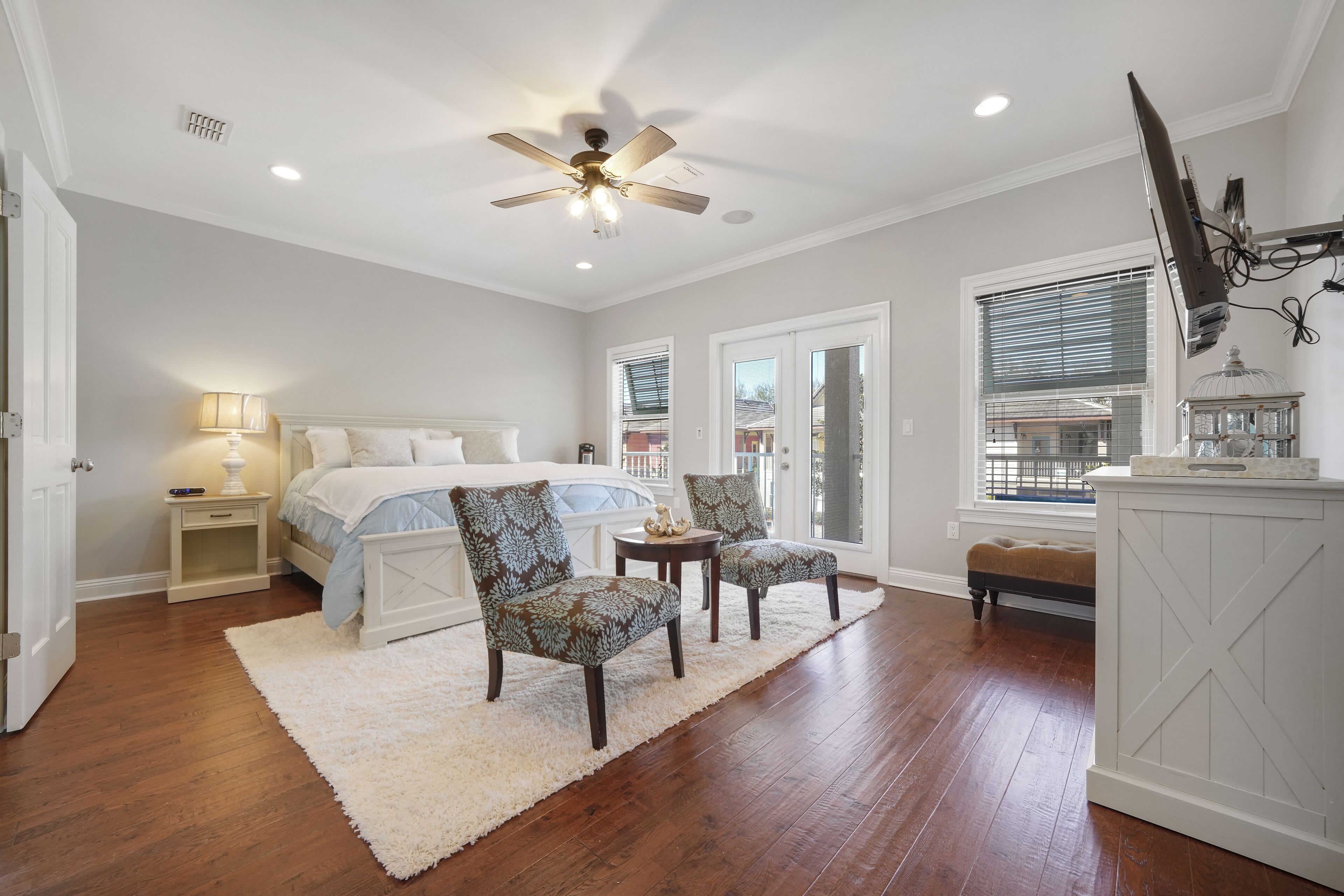 A bedroom with a bed, two patterned chairs, a white rug, a ceiling fan, and a large window with blinds.