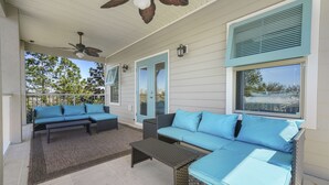 A covered outdoor seating area with wicker furniture and blue cushions, a ceiling fan, and a view of trees and a house in the background. - Beach Daze New 5BR, 4BA, 16 guest, Game Room. (Destin)