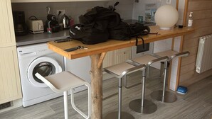 Une cuisine compacte équipée d’un lave-linge, d’un four à micro-ondes, d’un grille-pain et d’une machine à café. On y trouve une table en bois avec des tabourets, ainsi qu’un grand luminaire sphérique fixé au mur.