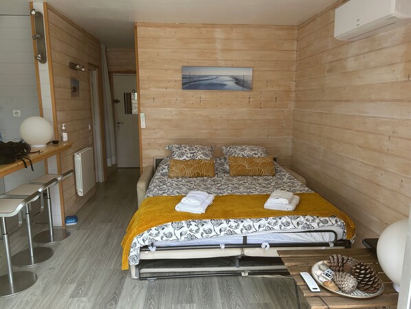 A compact room with a bed, a small table, a chair, and a wall-mounted air conditioner.