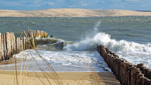 Un paysage côtier avec une jetée en bois menant à une plage de sable, des vagues se brisant contre la jetée et une dune en arrière-plan.