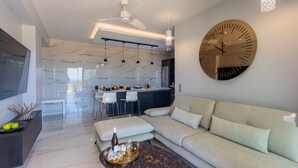 A modern living room with a sofa, a coffee table, a large wall clock, and a kitchen in the background. - AVITHOS BLUE VILLA  (kefalonia)
