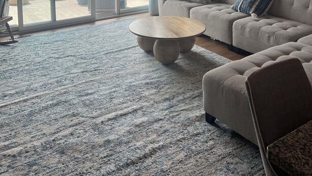 Ein geräumiges Wohnzimmer mit einer großen Schiebeglastür, die auf einen Balkon führt, einer grauen Sofagarnitur, einem runden Couchtisch und einer Kristallleuchtre.