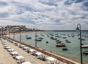 Un paseo marítimo con hileras de sillas y mesas de plástico blanco, una playa al fondo y numerosos barcos en el agua.