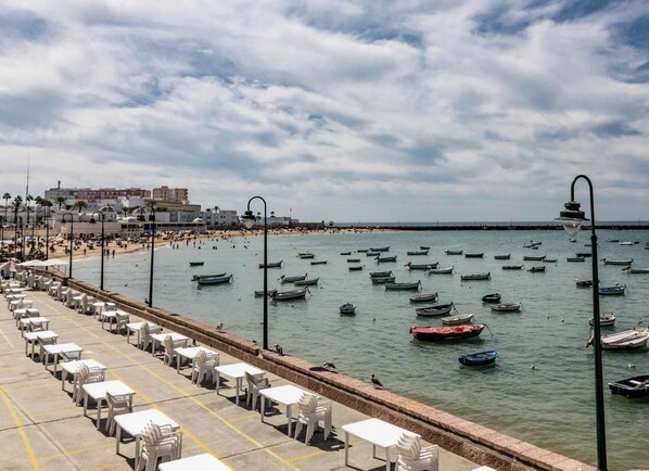 Une promenade en bord de mer bordée de rangées de chaises et de tables en plastique blanc, une plage en arrière-plan et de nombreux bateaux sur l’eau.