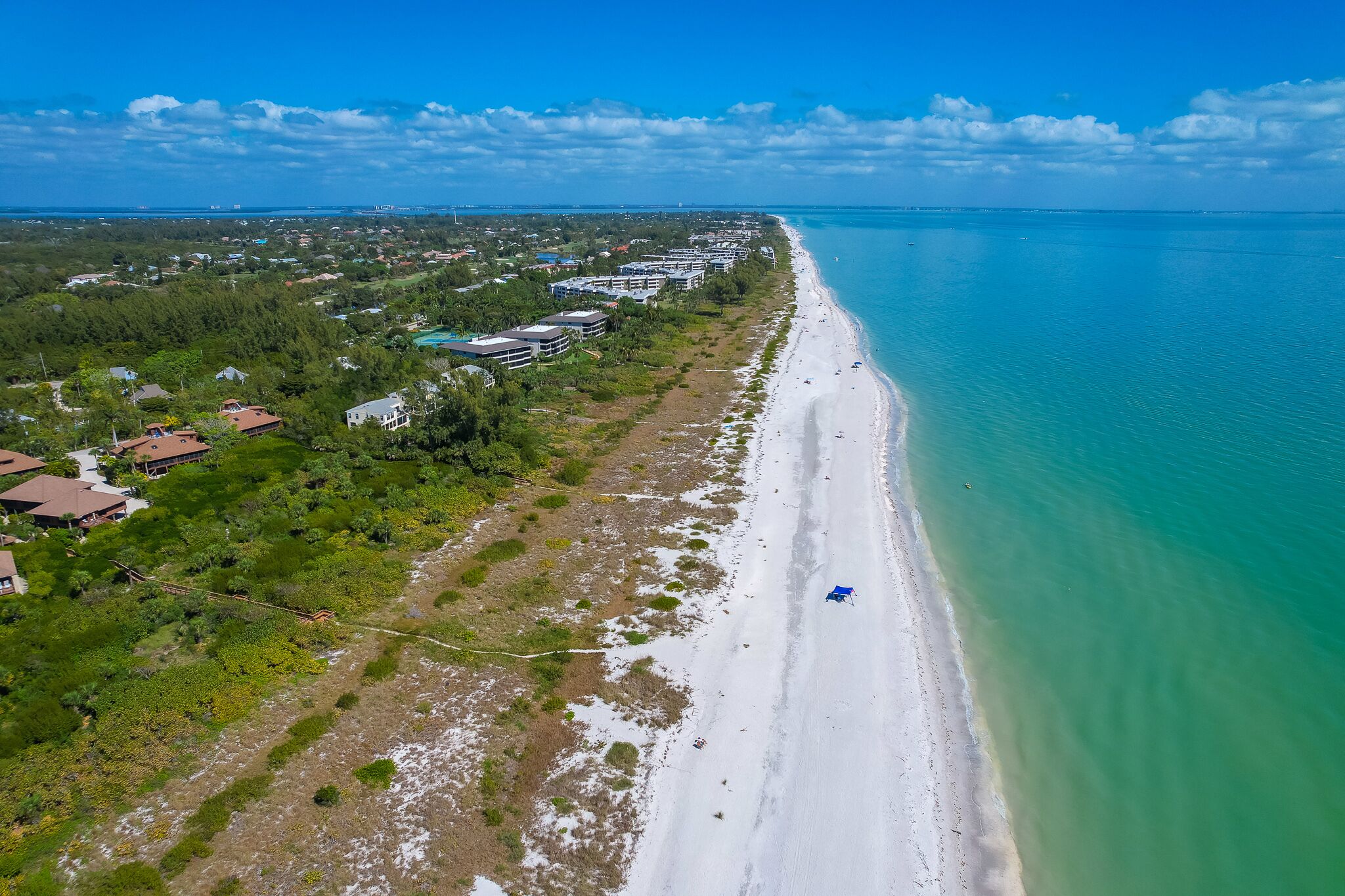 Eine Vogelperspektive einer Küstenlandschaft mit einem langen Sandstrand, einer Reihe von Gebäuden und einem klaren, blauen Himmel.
