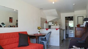 A compact living space with a red sofa, a dining table with chairs, a kitchen area with a microwave, and a small dining table with a vase of flowers. - Appartement T2 40 M² dans villa avec jardin Semaine /WE 2 personnes (Marseille)