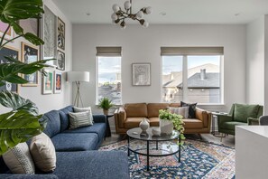 A modern living room with a blue sofa, a brown leather sofa, a glass coffee table, and a green armchair. There are two windows with blinds, a floor lamp, and a variety of framed artwork on the walls.