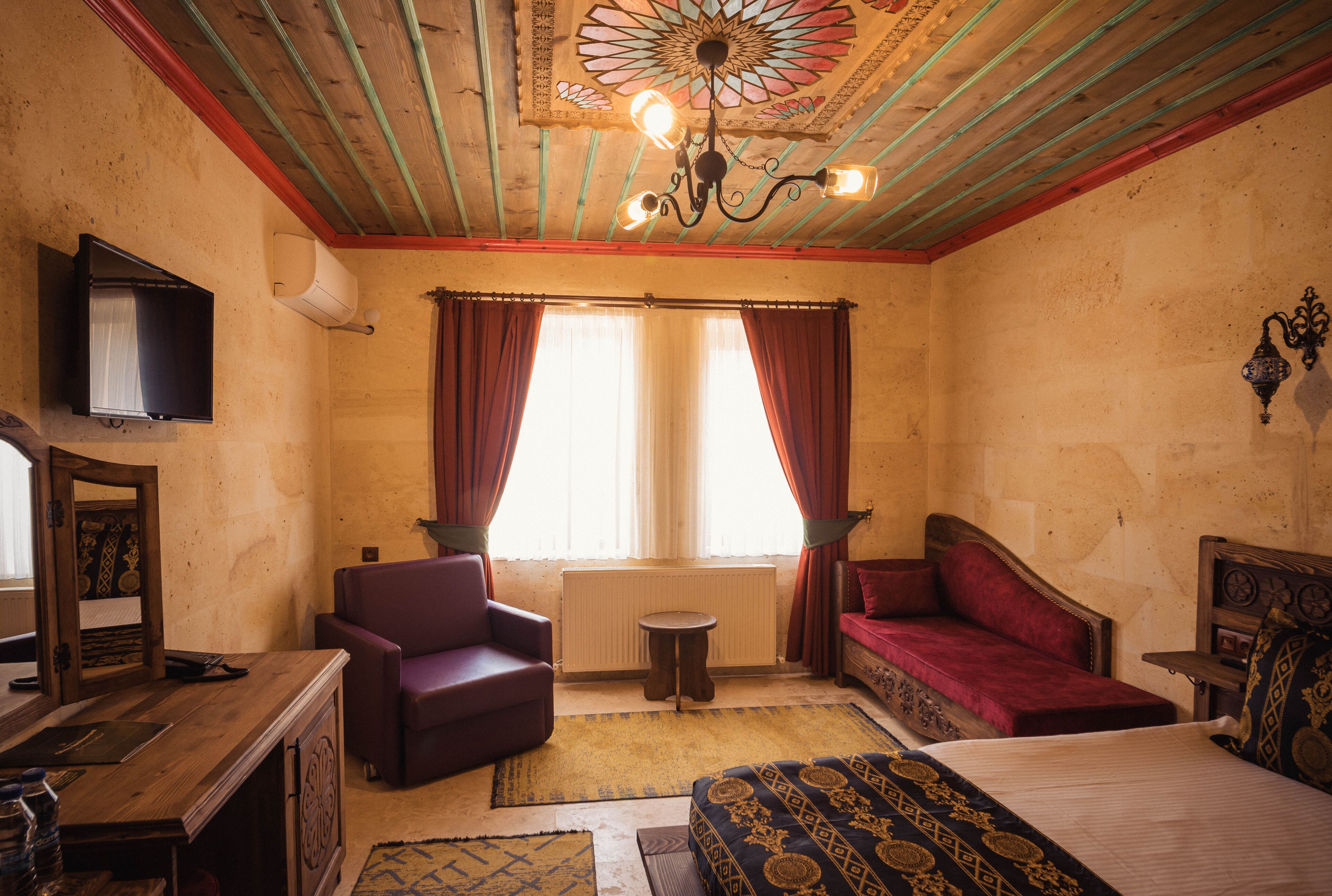 A room with a bed, a sofa, a chair, a small table, a television, and a decorative ceiling.